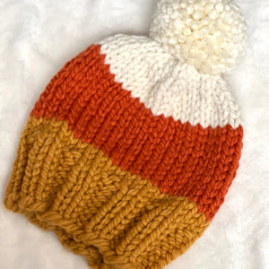 A hand-knit beanie hat with a chunky white pom-pom, featuring three horizontal color bands: white at the top, rust orange in the middle, and mustard yellow at the bottom, laid flat on a white textured fabric background.