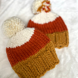 Two hand-knit baby hats styled like candy corn, featuring white, orange, and mustard yellow stripes with matching pom-poms, laid on a white fabric background.