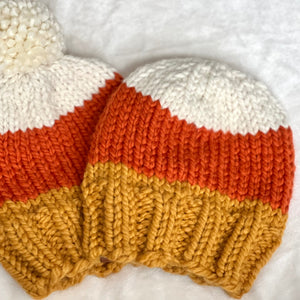 Two hand-knit baby beanies with a white pom-pom, featuring horizontal stripes of white, orange, and mustard yellow, displayed on a white textured background.