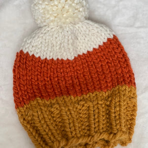 A hand-knit beanie hat with a white pom-pom, featuring horizontal stripes of white, burnt orange, and mustard yellow, laid flat on a white textured background.