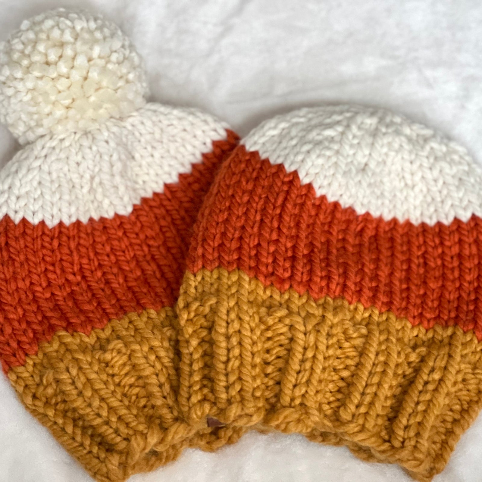 Two hand-knit baby beanies with a white pom-pom, featuring horizontal stripes of white, orange, and mustard yellow, laid flat on a white textured blanket.