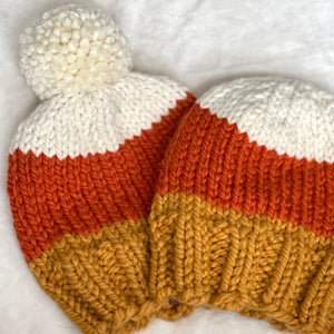 Two hand-knit baby beanies with a white pom-pom, featuring horizontal stripes of white, burnt orange, and mustard yellow, displayed on a soft white textured fabric background.