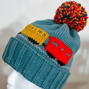 A teal knit beanie with a multicolor pom-pom and a train motif in yellow, red, black, and gray, displayed on a white mannequin head.