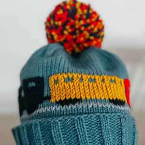 A close-up of a teal knit beanie with a multicolor pom-pom on top, featuring a yellow, black, and gray knit pattern band around the upper portion.