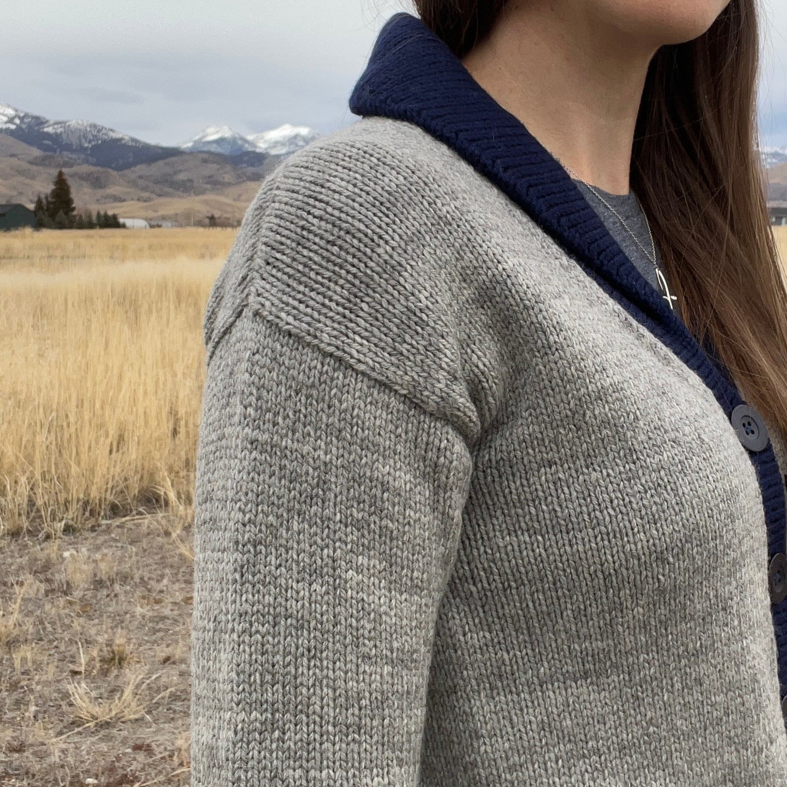 Person wearing a beige and brown cardigan in a field with mountains in the background