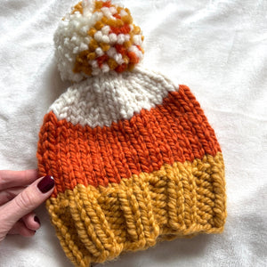 A hand-knit baby beanie hat styled like a candy corn, featuring white, orange, and mustard yellow stripes with a matching pom-pom, held by a hand on a white textured fabric background.