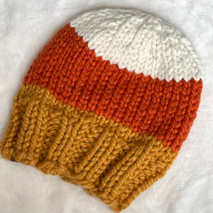 A chunky hand-knit beanie hat with a three-color candy corn design, featuring white on top, orange in the middle, and mustard yellow on the bottom, laid flat on a white textured fabric background.