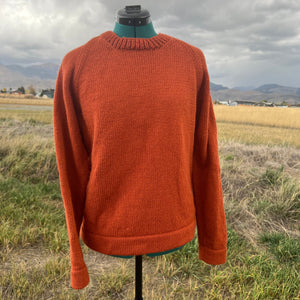 An orange knit crewneck sweater displayed on a mannequin torso outdoors in a grassy field under a cloudy sky.