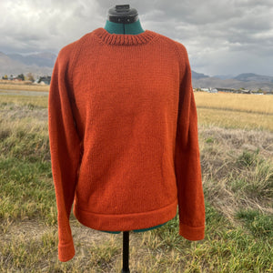 An orange knit crewneck sweater displayed on a dress form mannequin outdoors, with a blurred field and mountains in the background.