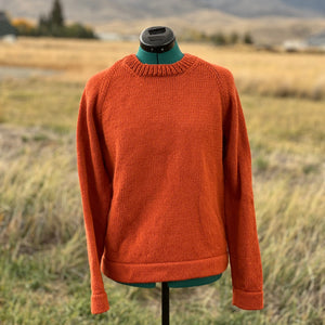 An orange knit crewneck sweater displayed on a mannequin torso stand in an outdoor grassy field with mountains in the background.