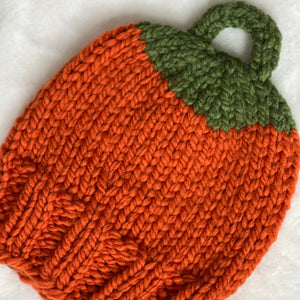 A hand-knit pumpkin-shaped baby hat or bootie in bright orange yarn with a green stem and loop handle, laid flat on a white textured fabric background.