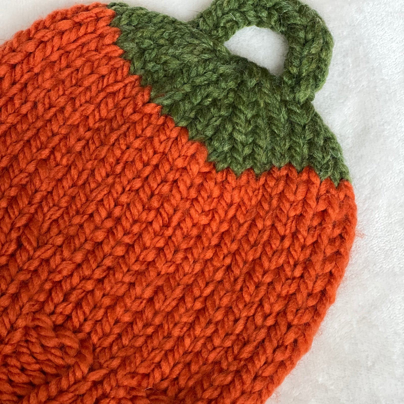 A hand-knit pumpkin-shaped beanie hat in bright orange with a green leafy top, laid flat on a white textured fabric background.