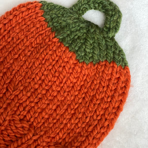 A close-up of a chunky knit pumpkin-shaped item, likely a cozy or pouch, featuring a vibrant orange body and a green stem detail, laid flat on a white textured fabric background.