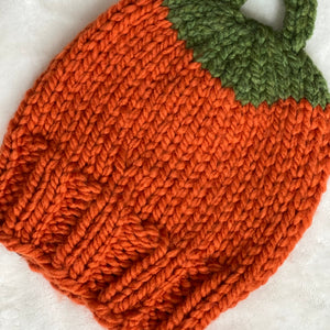 A close-up of a hand-knit pumpkin-shaped item, likely a pouch or small bag, featuring a textured orange body and a green leafy top, laid on a white fabric background.