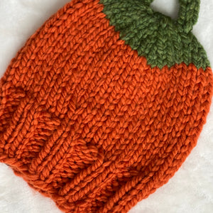 A hand-knit pumpkin-shaped baby hat or bootie in bright orange yarn with a green stem and loop handle, laid flat on a white textured fabric background.
