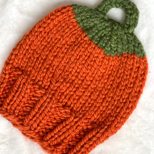 A hand-knit pumpkin-shaped beanie hat in bright orange with a green leafy top, laid flat on a white textured fabric background.