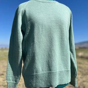 A mint green knit sweater displayed on a dress form mannequin outdoors against a blue sky and dry grassy field background.
