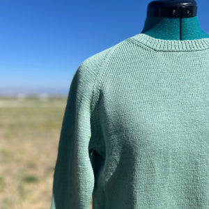 A mint green knit sweater displayed on a black dress form mannequin outdoors, with a blurred grassy field and blue sky in the background.