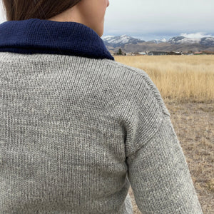 Person wearing a gray sweater with a blue collar standing in a field with mountains in the background