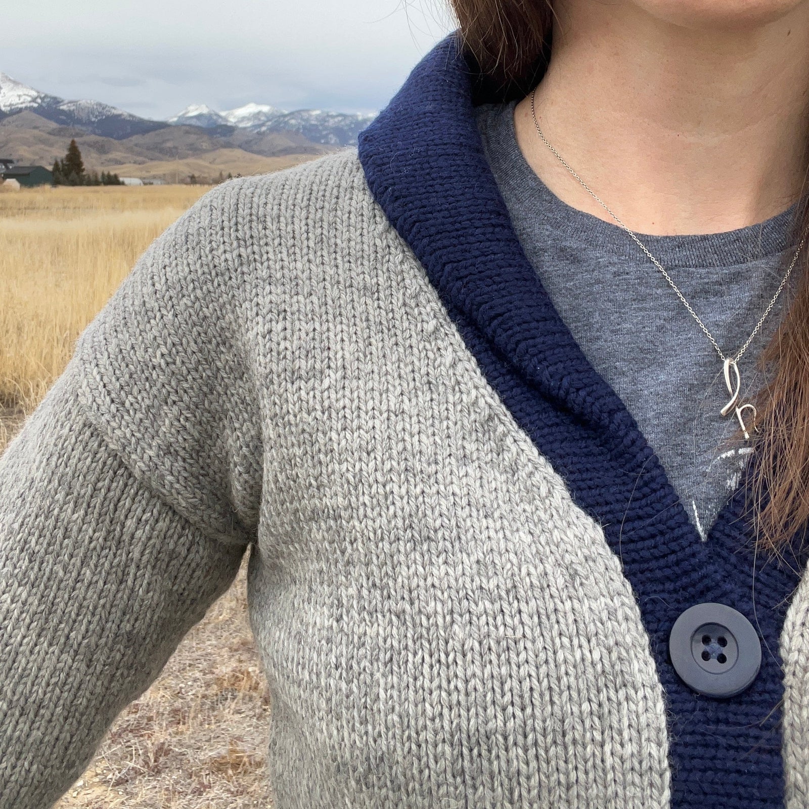 Person wearing a gray cardigan with navy trim in a field with mountains in the background