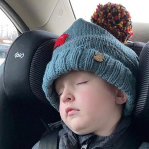 A sleeping child wearing a teal knit beanie with a red heart and a multicolor pom-pom, seated in a car seat.
