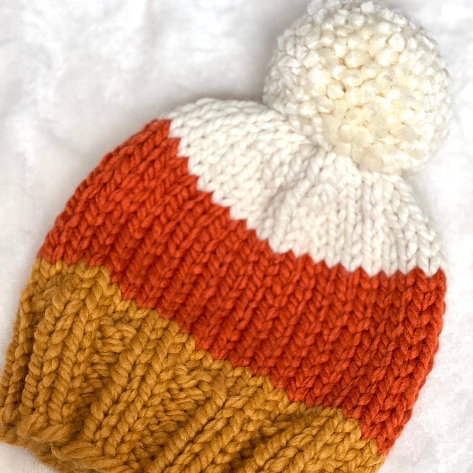 Two hand-knit baby beanies with a white pom-pom, featuring horizontal stripes of white, orange, and mustard yellow, laid flat on a white textured blanket.