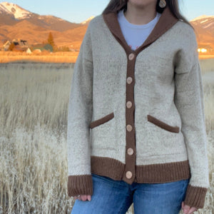 Person wearing a beige and brown cardigan in a field with mountains in the background