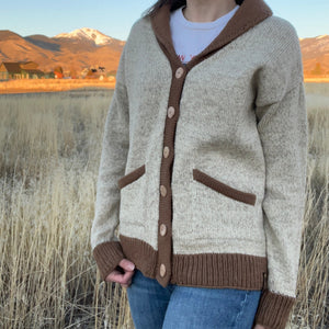 Person wearing a beige and brown cardigan in a field with mountains in the background