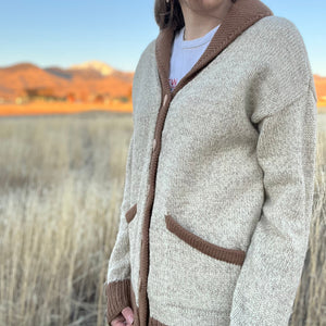 Person wearing a beige and brown cardigan with a scenic background of mountains and fields.
