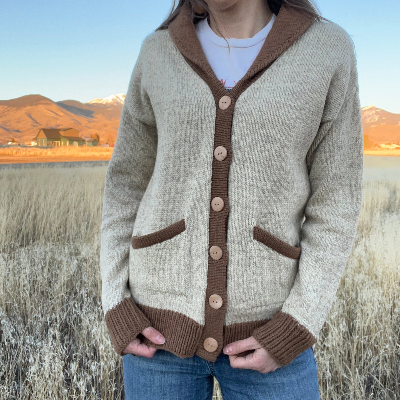 Person wearing a beige and brown cardigan in a field with mountains in the background