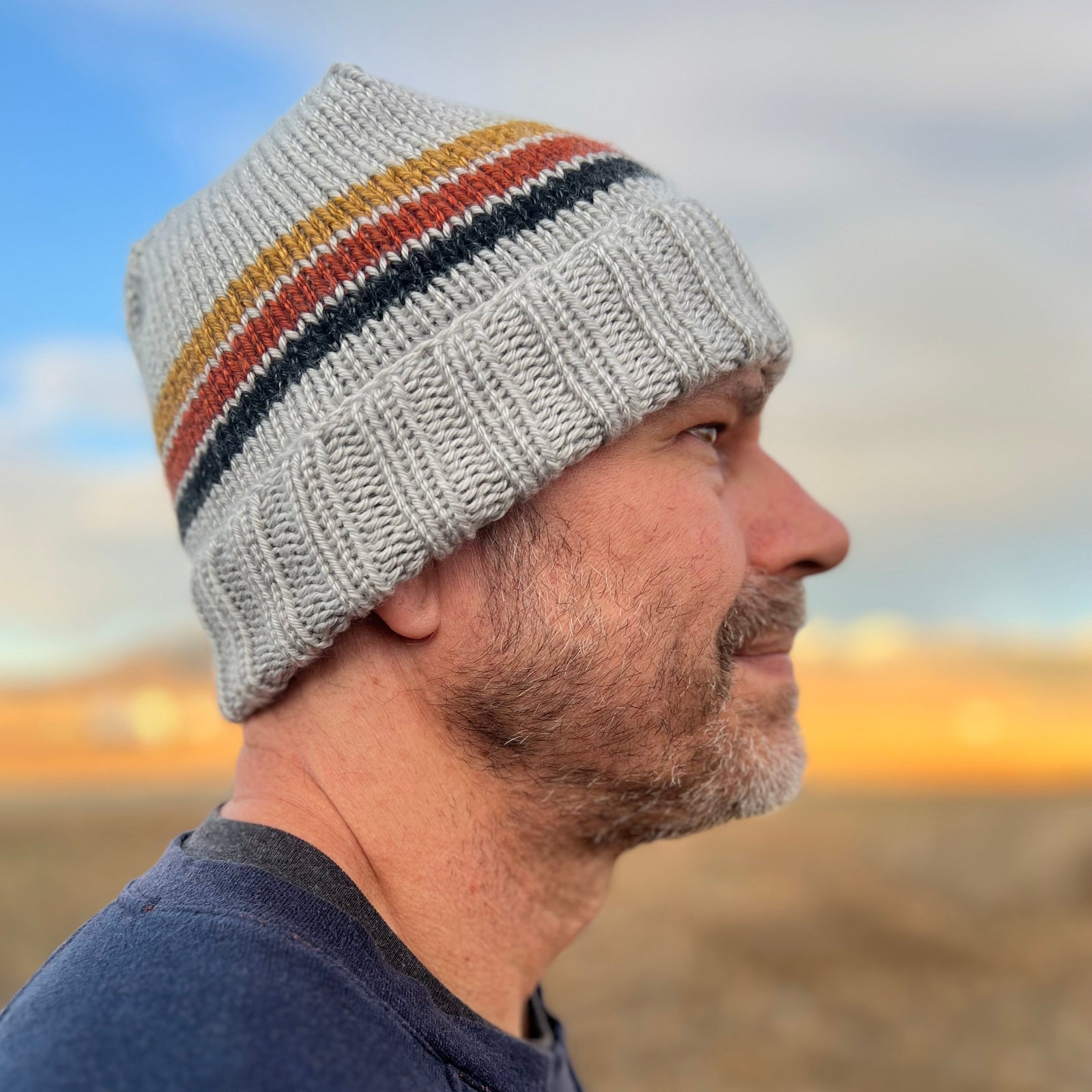 A person wearing a gray beanie with a striped pattern in white, yellow, and blue colors, looking off to the distance.