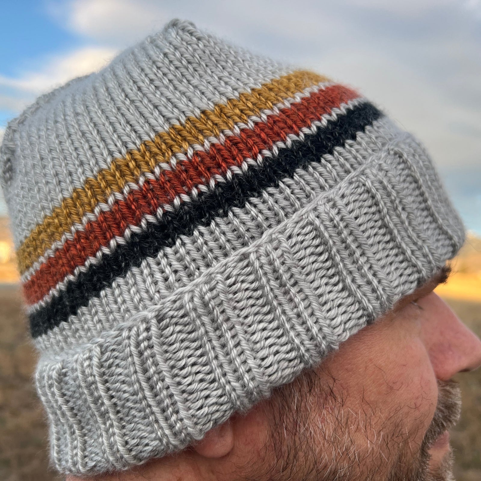 A person wearing a gray beanie with a striped pattern in white, yellow, and blue colors, looking off to the distance.