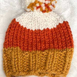 A chunky hand-knit beanie hat with a white top section, a wide orange middle section, and a textured mustard yellow lower section, finished with a matching pom-pom in orange, white, and mustard yellow, laid flat on a white textured background.