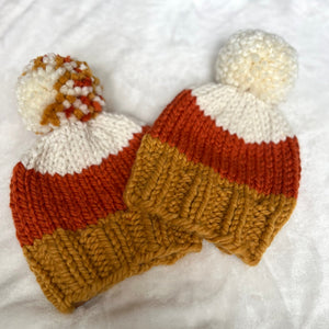 Two hand-knit baby hats in a candy corn color scheme of white, orange, and mustard yellow, each topped with a white pom-pom featuring orange and yellow yarn accents, displayed on a white textured fabric background.