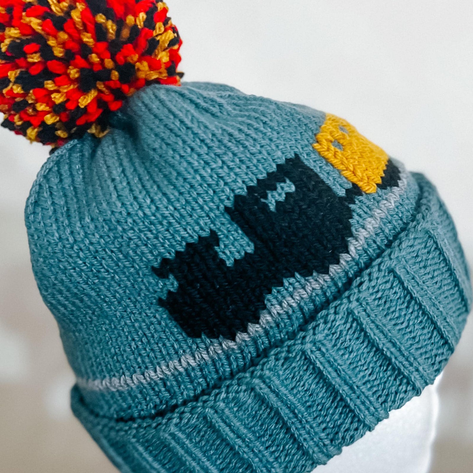 A close-up of a teal knit beanie hat with a multicolor pom-pom in red, orange, yellow, and black. The beanie features a dark navy blue pixelated graphic and a yellow accent on the side.