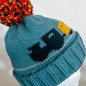 A close-up of a teal knit beanie hat with a multicolor pom-pom in red, orange, yellow, and black. The beanie features a dark navy blue pixelated graphic and a yellow accent on the side.