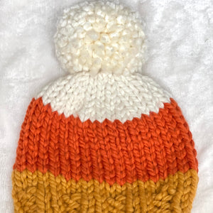 A hand-knit beanie hat with a fluffy pom-pom, featuring three horizontal stripes in cream, orange, and mustard yellow, laid flat on a white textured fabric background.
